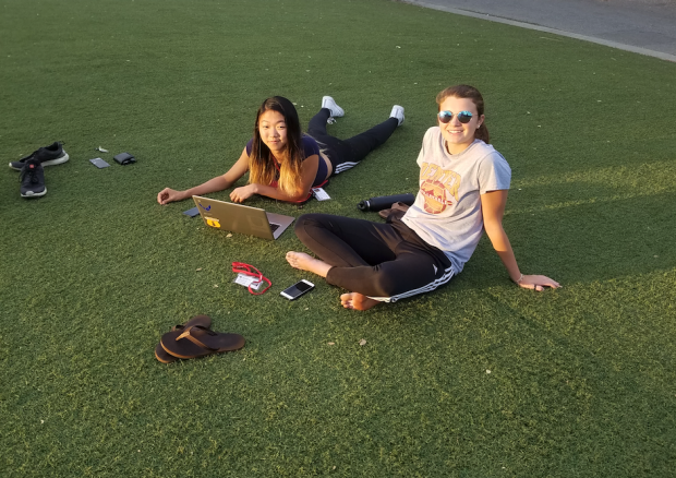 Two girls sit on field with open laptop