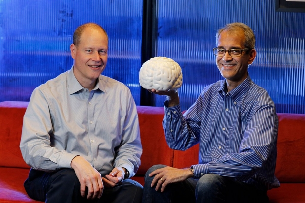 Two men sitting on a couch with one of them holding up a model of a brain